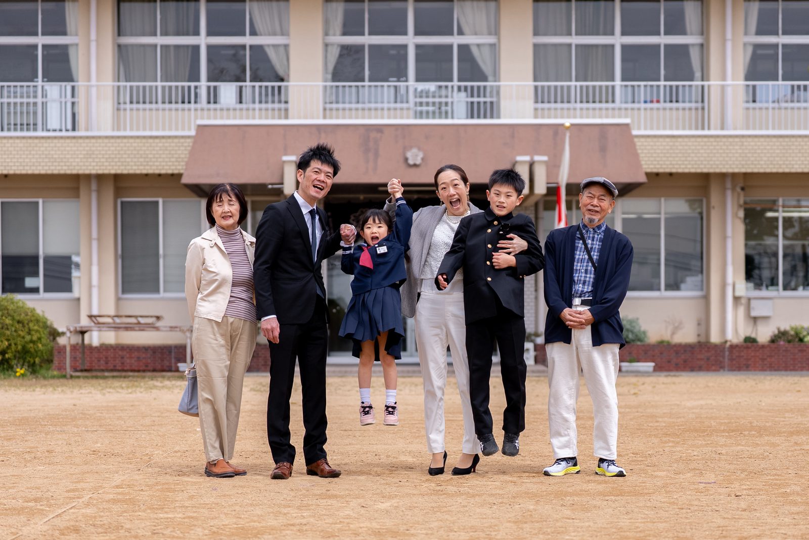 校舎を背景に、祖父母と両親、兄妹の6人で手を上げて跳ぶ集合写真
