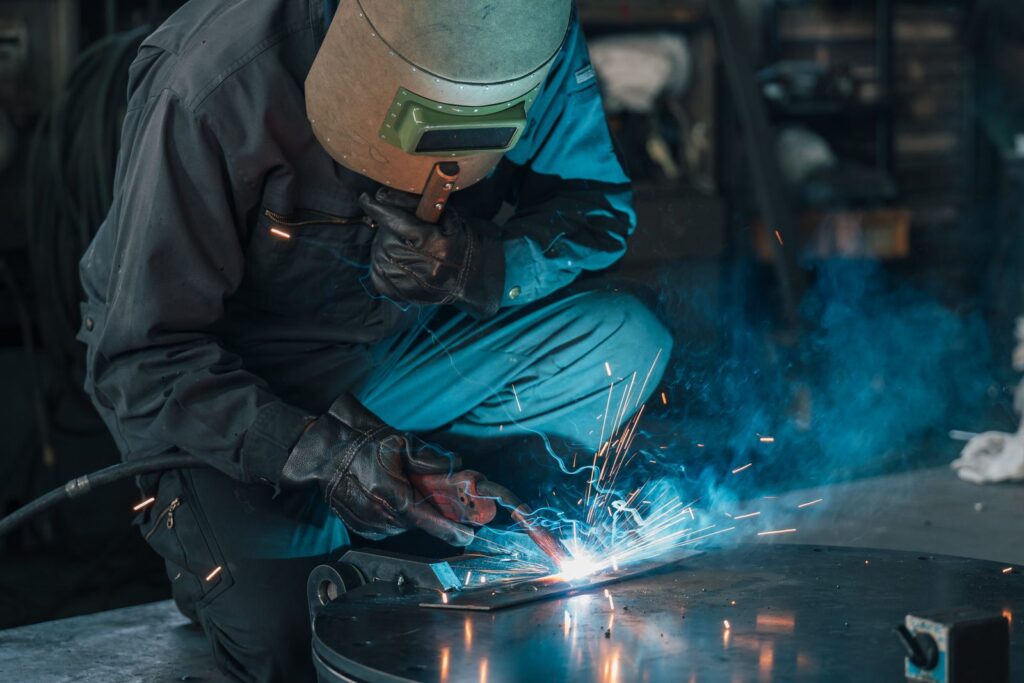 火花と煙のなかで溶接する職人。青い作業着と保護面